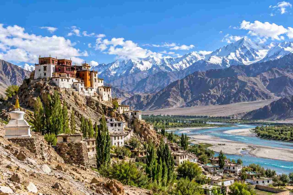 Traditional Buddhist monastery overlooking a turquoise river valley and snow-capped Himalayan mountains in Ladakh, India.